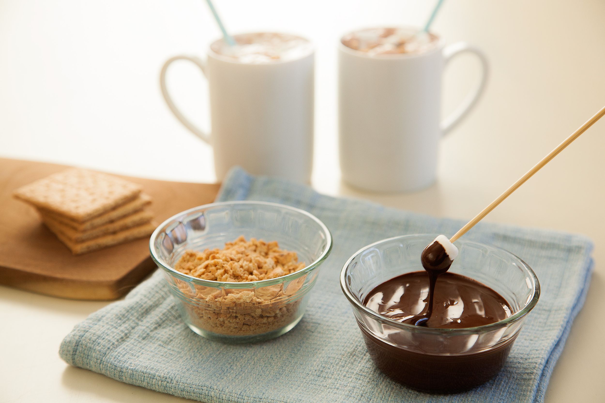 Two mugs of hot cocoa beside chocolate sauce and crushed graham crackers